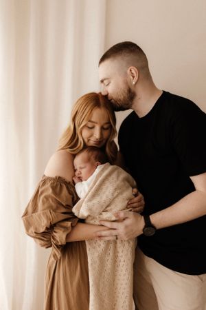 Family portrait in neutral tones against white wall with sleeping newborn.