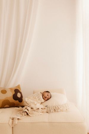 Sleeping baby wrapped in cream colored blanket rests peacefully on white couch with decorative pillow.
