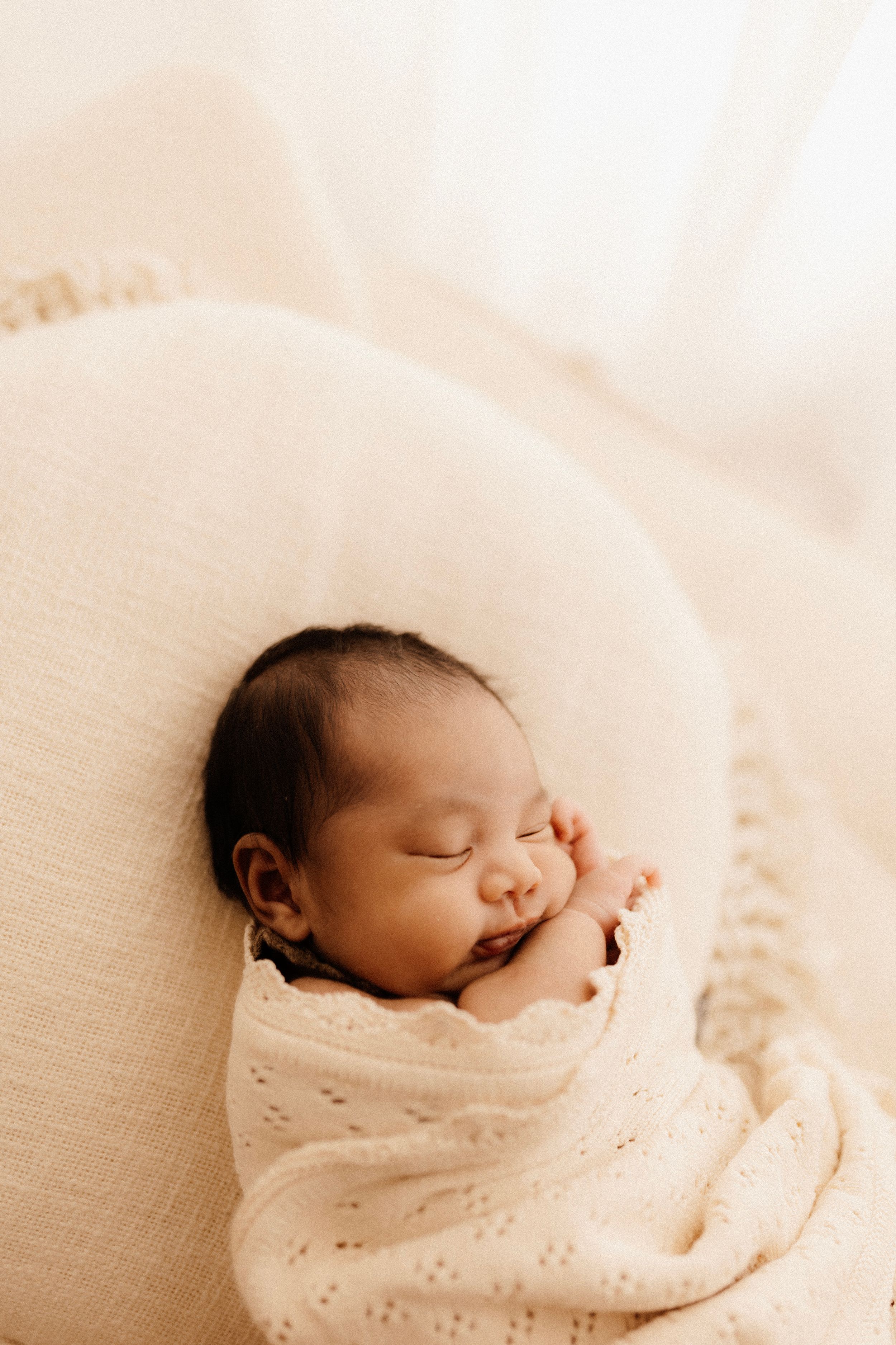 A sleeping newborn wrapped in cream blanket yawns while nestled in white cushions.