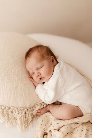Sleeping newborn in white onesie resting on textured cream pillow.