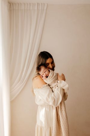 A tender moment captured in soft light as parent holds newborn baby against white curtain backdrop.