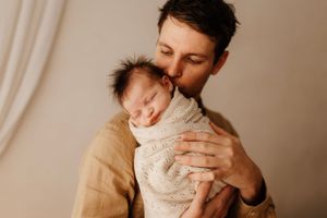 Parent tenderly holding and kissing sleeping newborn swaddled in cream fabric.