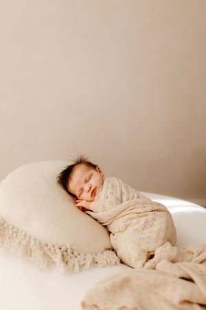 Sleeping newborn with dark hair wrapped in cream knit blanket resting on beige pillow.