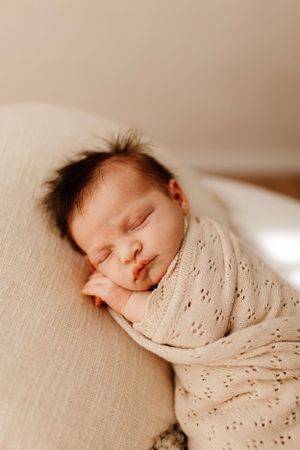 Close up of peaceful sleeping newborn wrapped in textured cream blanket on beige cushion.
