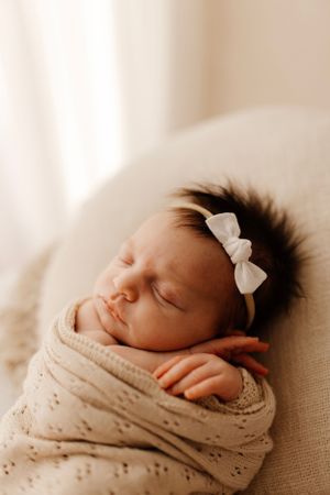 Sleeping baby with white bow headband wrapped in cream knit blanket on cushion.