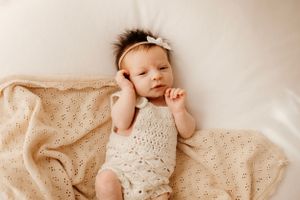 Baby wearing lace outfit and bow headband lying on cream textured blanket.