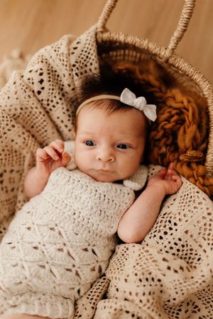 Baby in cream knit outfit with bow headband lying in woven basket with textured blanket.