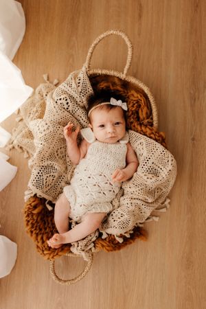 Overhead view of baby in cream knit dress lying in woven basket with decorative blanket.