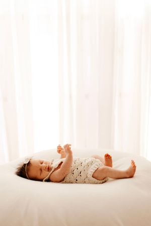 Baby wearing lace outfit lying peacefully on white bedding in soft light.