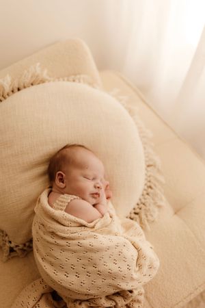 Sleeping newborn wrapped in cream knit blanket resting on beige cushion.