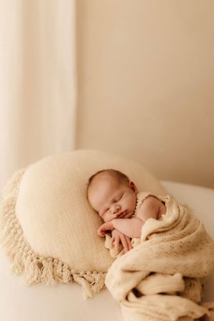 Peaceful newborn sleeping on cream pillow wrapped in knitted blanket.