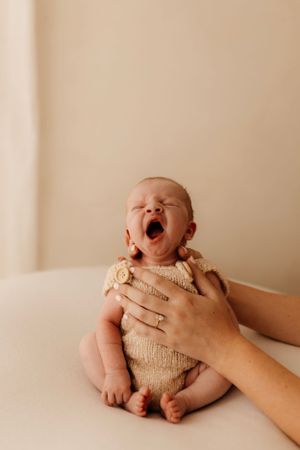 Yawning baby in knit outfit sitting supported by hands against light background.