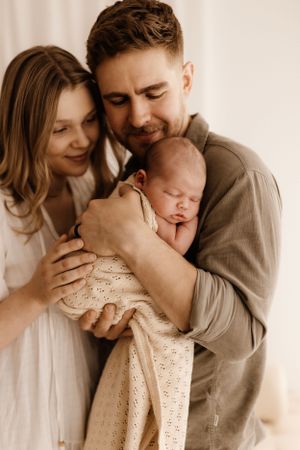 A tender family moment as parents cradle their sleeping newborn in warm neutral tones.