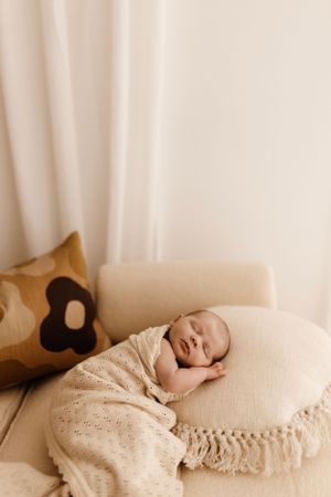 A sleeping newborn wrapped in beige blanket rests on cream pillow beside decorative cushion.