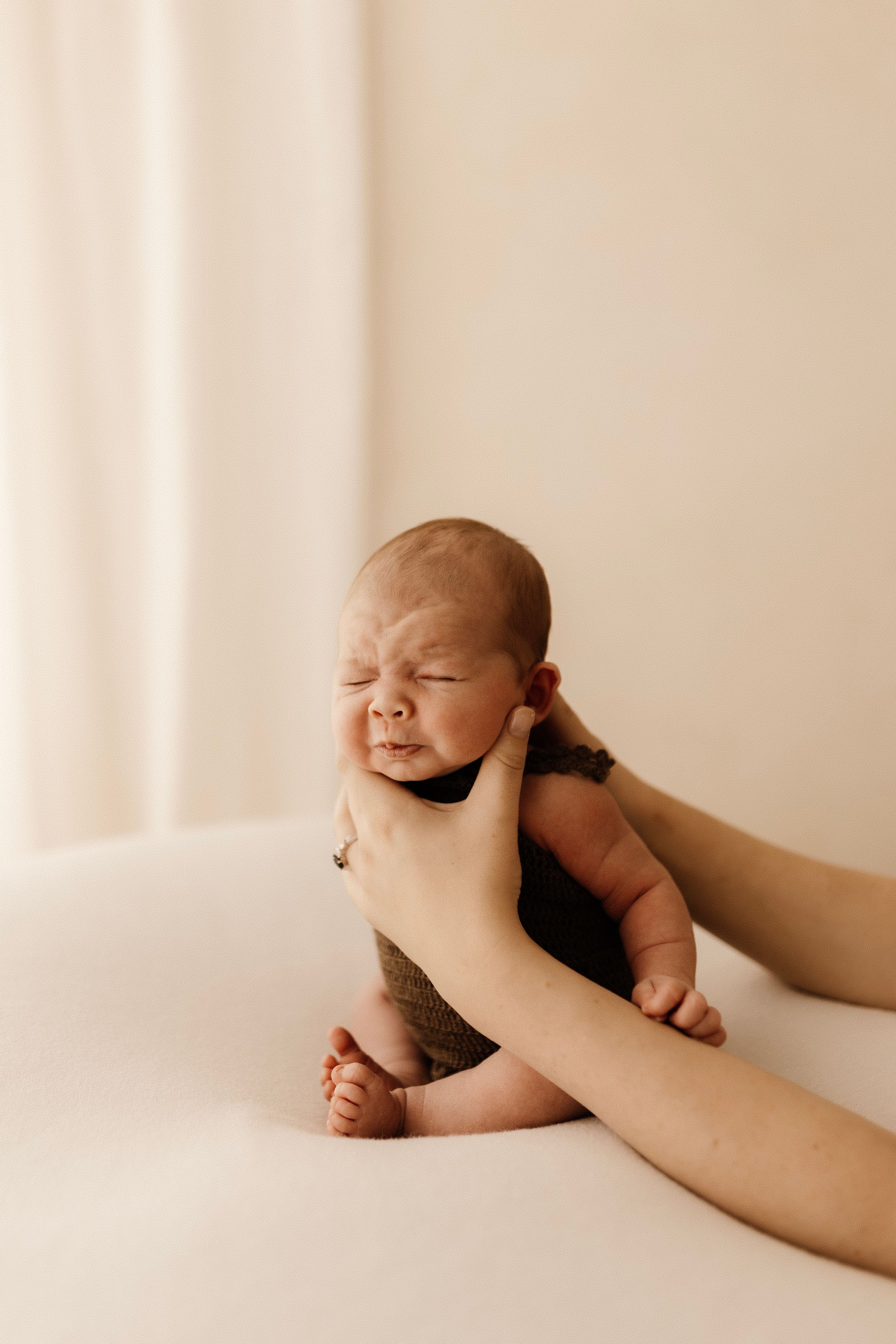 A baby wearing a brown outfit expresses emotion while being supported by gentle hands.
