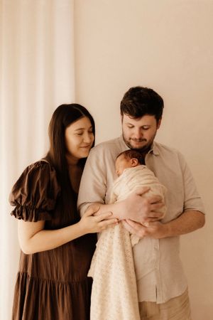A tender family moment as parents hold their swaddled newborn against neutral backdrop.