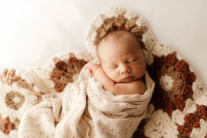 Sleeping newborn wrapped in cream blanket with floral crown on textured brown blanket.