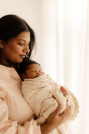 Mother cradles sleeping newborn wrapped in knit blanket by window.
