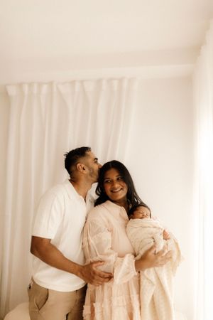 A tender family moment captured in soft lighting against white curtains as parents embrace their newborn baby.