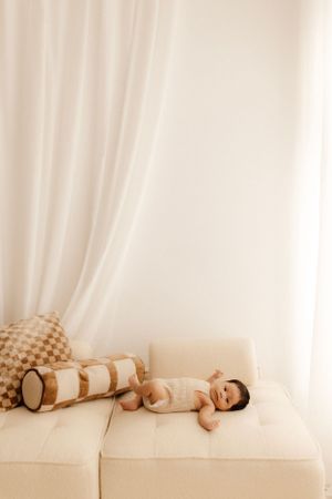 A peaceful newborn lying on a cream-colored couch with decorative pillows in natural light.