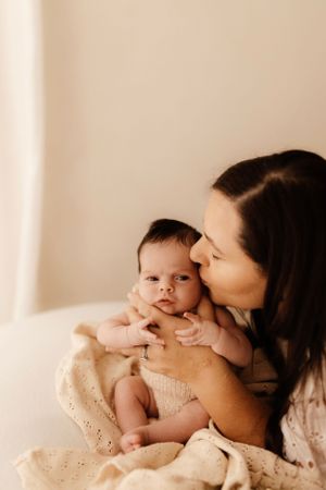 Mother in white lace dress kisses her baby while holding them in warm natural light.