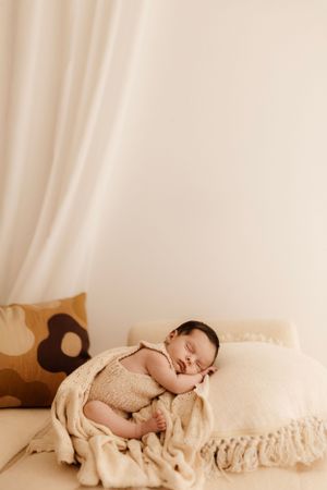 A sleeping newborn wrapped in beige fabric rests peacefully on a couch with earth-toned pillows.