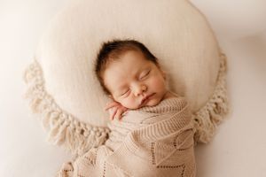 Sleeping baby in knit sweater nestled on cream pillow with fringe detail.