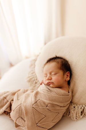 Peaceful newborn sleeping on white cushion wearing beige knitted outfit.