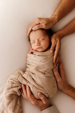 Hands gently cradle sleeping newborn wrapped in beige knit blanket against white background.