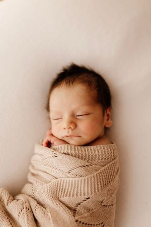 Sleeping newborn baby wrapped in cozy beige knit blanket against light background.