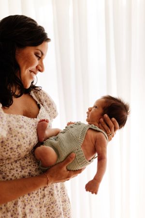Tender moment between parent and baby dressed in knit romper by window with white curtains.