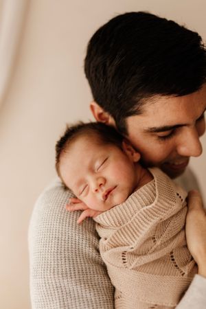 Peaceful sleeping newborn in beige knit sweater rests against shoulder in gentle embrace.