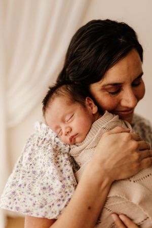 Newborn baby in floral dress sleeps peacefully against parent wearing beige sweater in soft lighting.