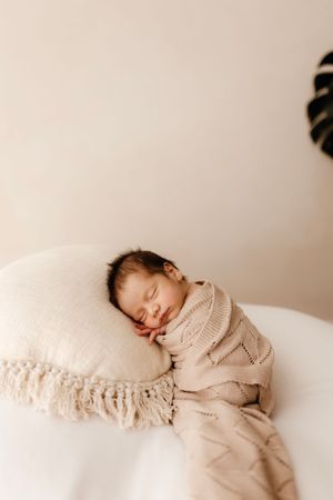 Sleeping newborn in beige knit wrap rests on white fringed pillow.