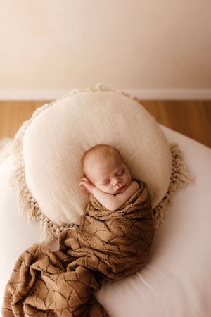 Sleeping newborn wrapped in brown knit blanket on round white cushion.
