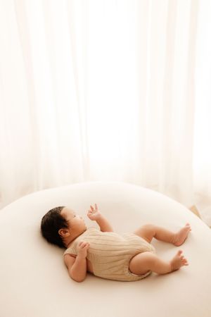 Baby in cream-colored outfit lying peacefully on white curved surface.