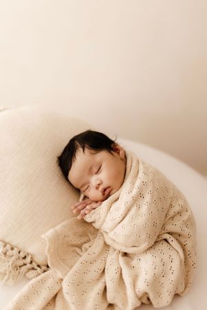 Peaceful sleeping baby wrapped in cream eyelet fabric resting on soft pillow.