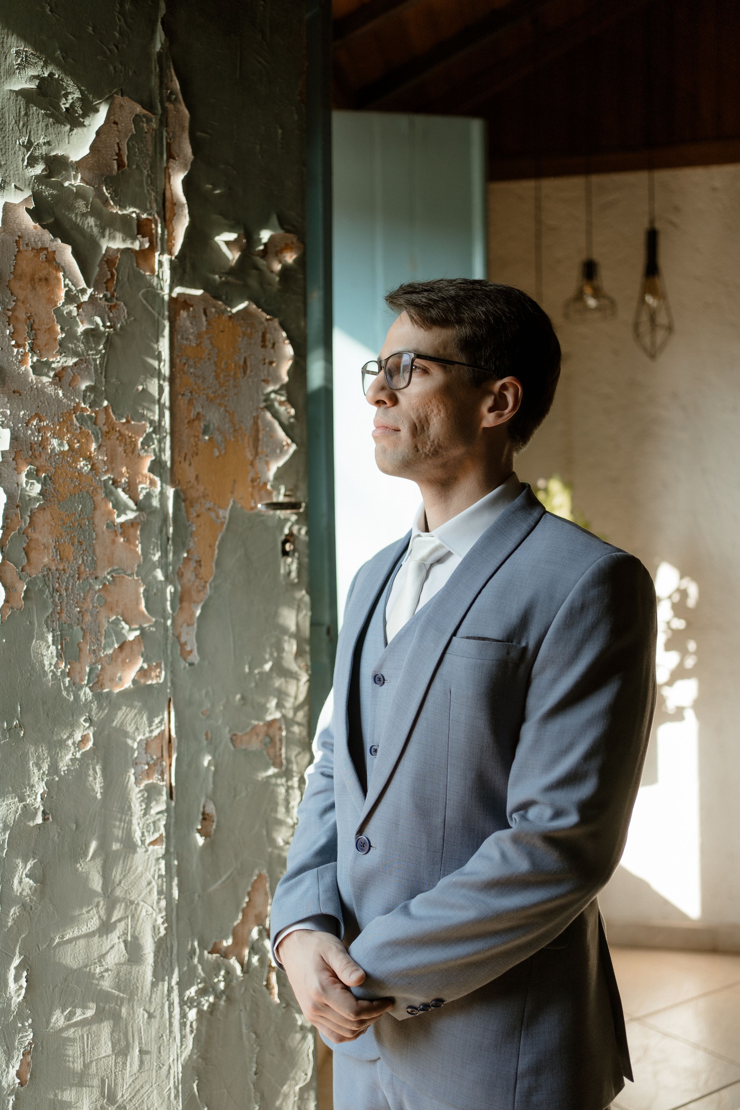 A suited figure stands thoughtfully by a weathered wall in dramatic lighting.