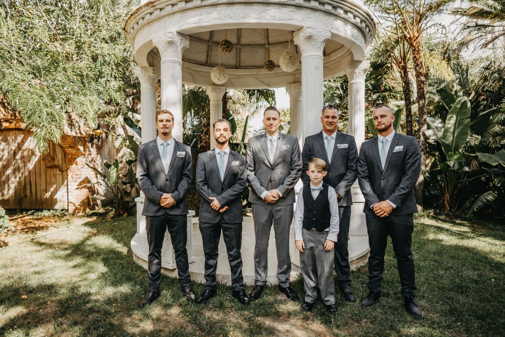 Groomsmen in gray suits pose together under stone gazebo in garden setting.