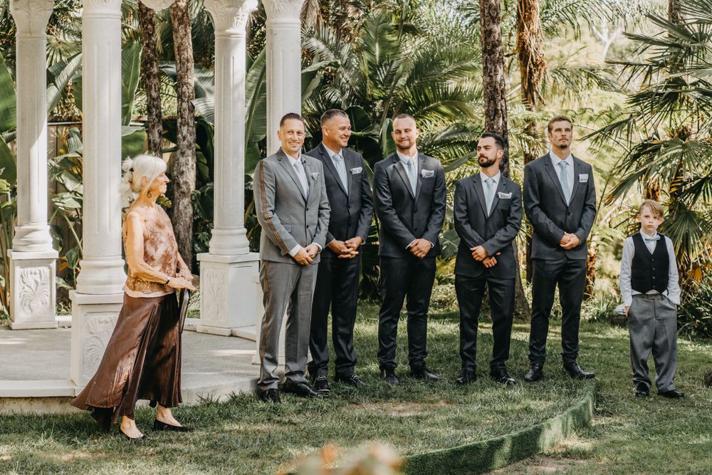 Wedding party lines up in formal attire next to white gazebo in garden setting.