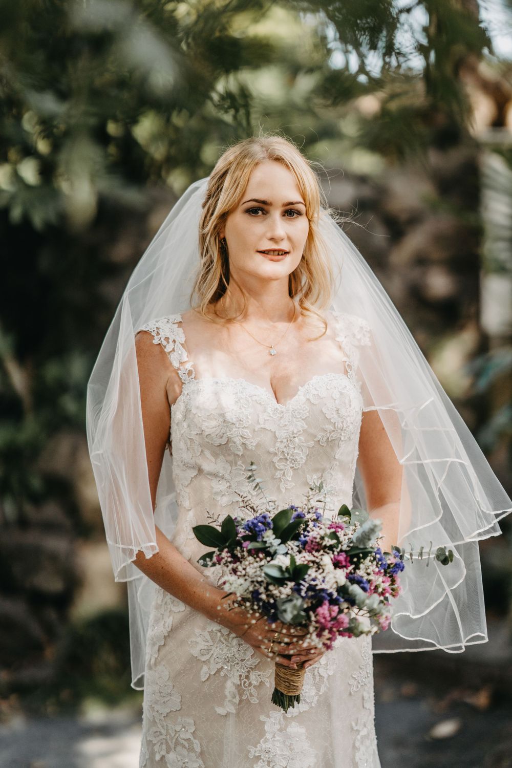 Bride in lace dress holds colorful bouquet while posing in garden setting.