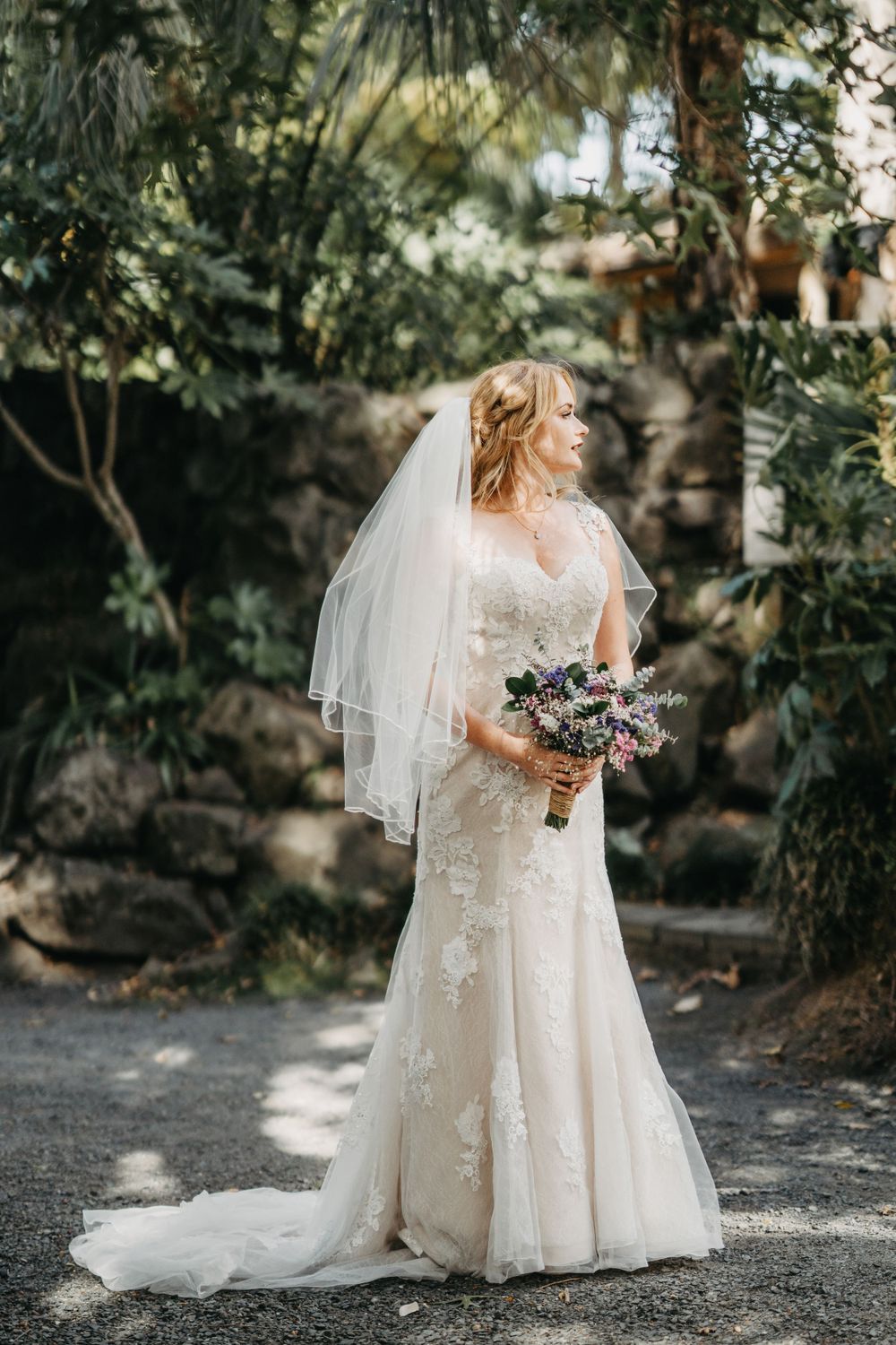 Elegant bride in lace wedding dress with flowing veil poses in garden setting holding purple and white bouquet.