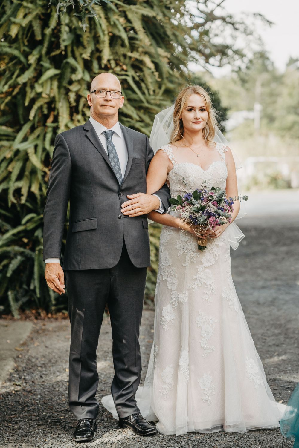 Wedding couple poses outdoors with bride in lace dress and groom in grey suit with purple bouquet.