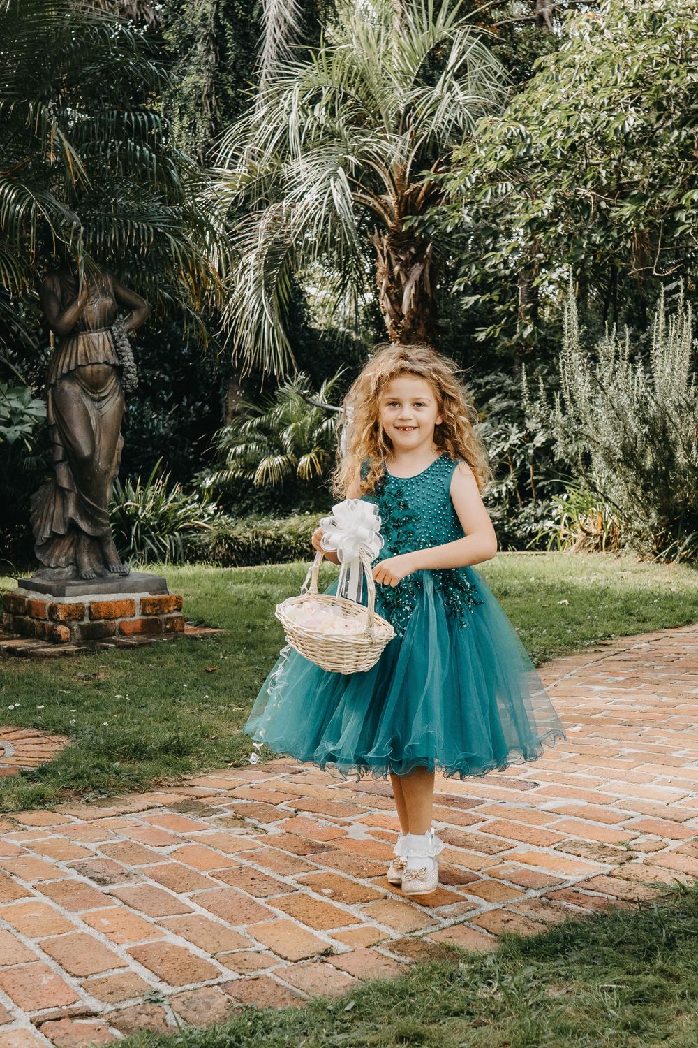 Young flower girl in teal tulle dress carries basket while walking down brick pathway.