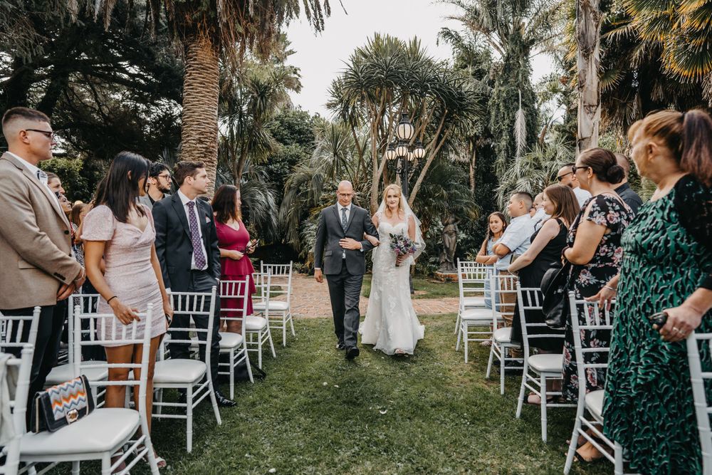 Wedding ceremony processional on outdoor garden path lined with white chairs and palm trees.