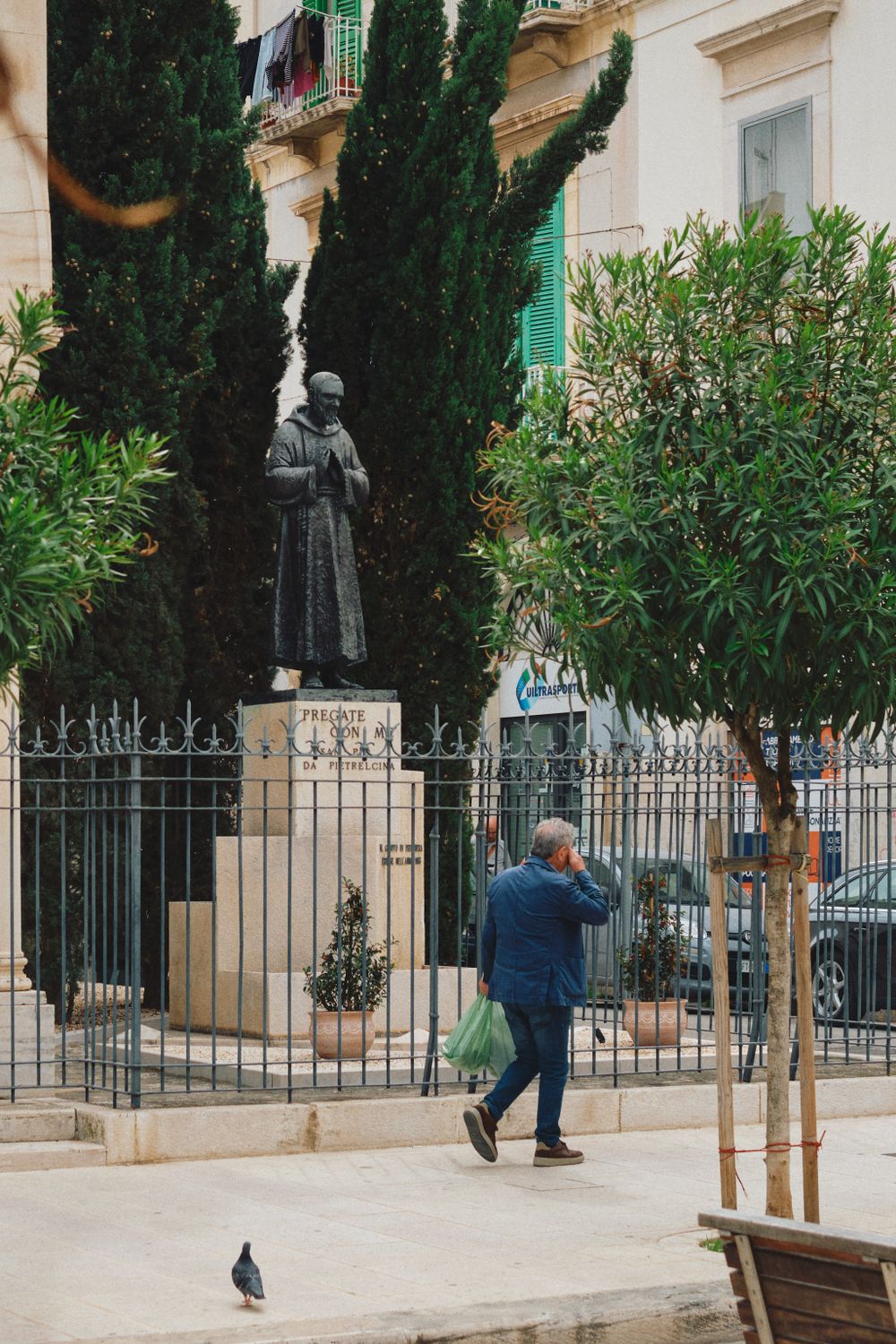 A religious statue stands among cypress trees and Mediterranean plants behind an iron fence.
