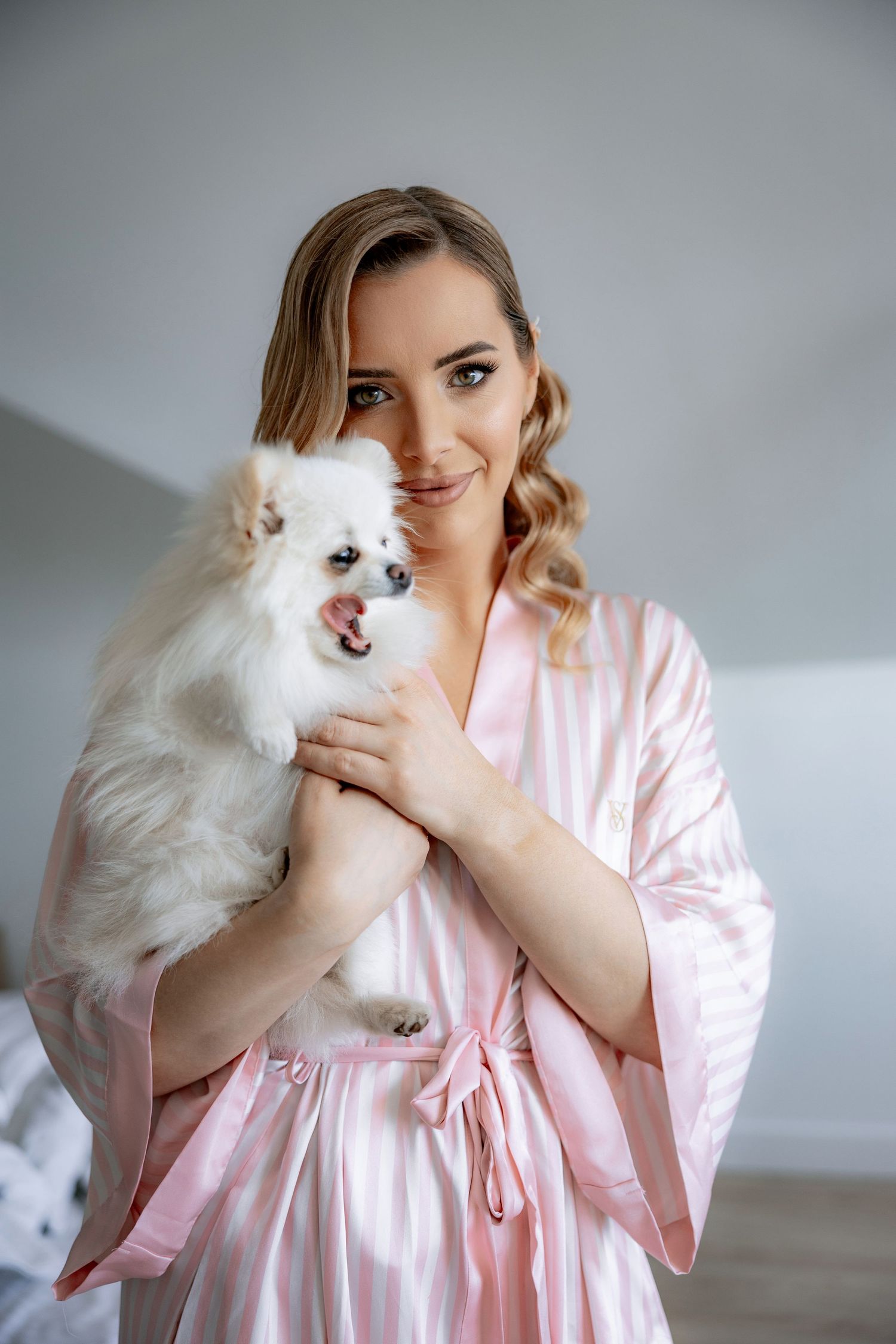 A fluffy white Pomeranian dog being held while wearing a pink satin robe.