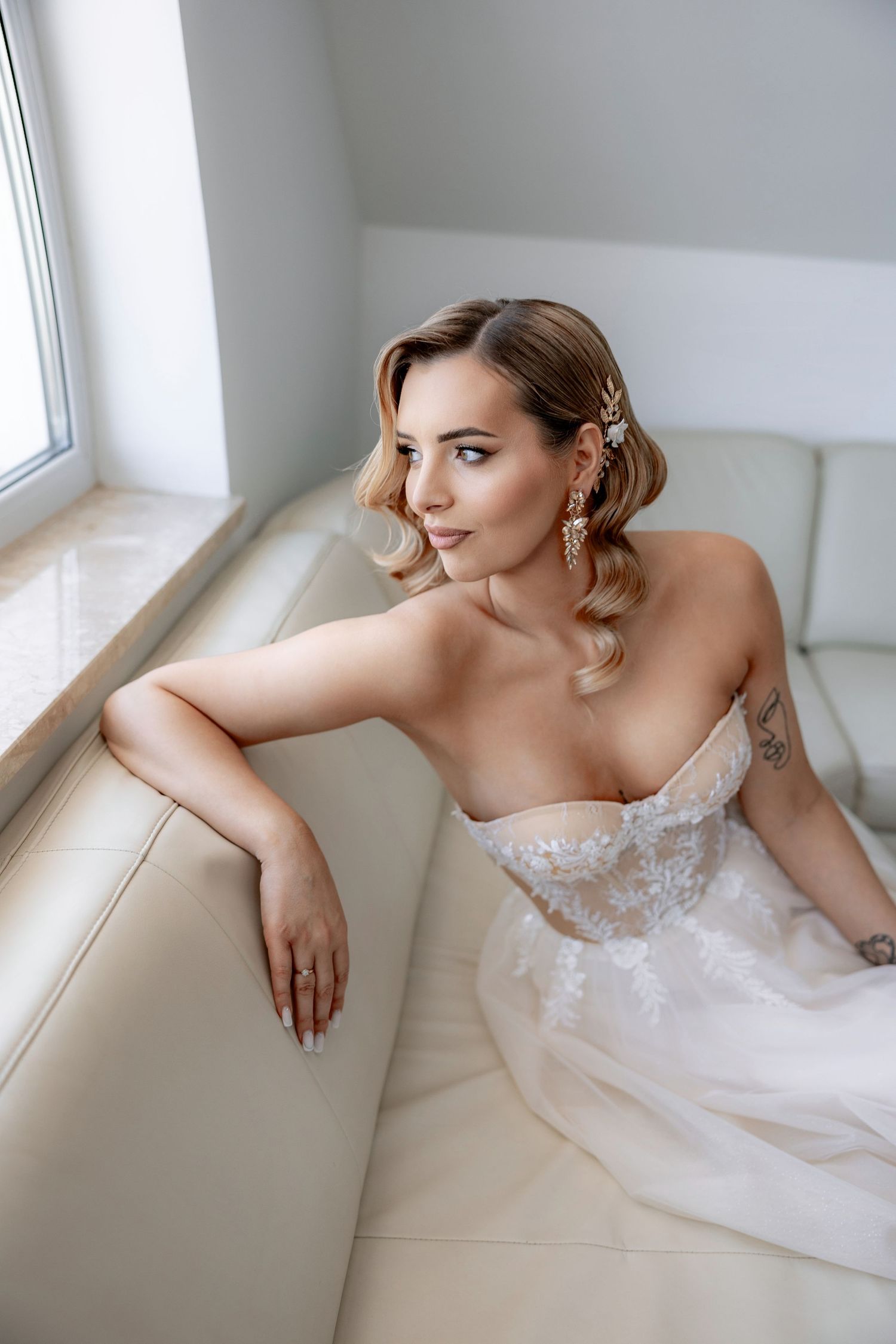 Bride in strapless lace wedding gown reclining on white leather couch near window.
