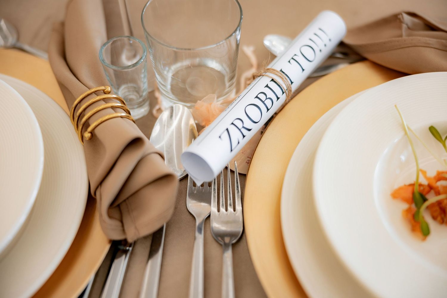 Wedding table setting with rolled napkin, white plates, and place card in neutral tones.