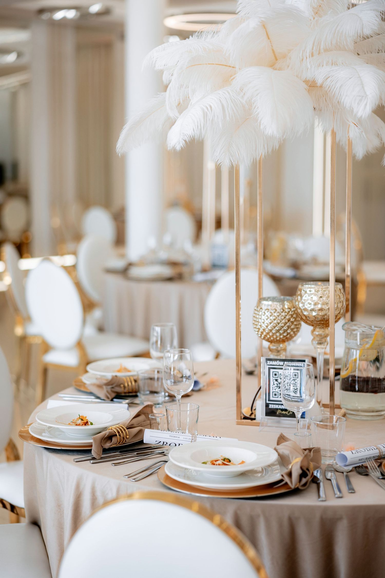 Elegant wedding reception table setting with white feather centerpieces, gold accents, and classic white dinnerware.